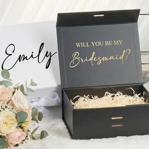 May include: Black gift box with gold text reading "Will you be my Bridesmaid?". A white box with "Emily" in black script is on the left. A white box with "Sophia" is on the right. Boxes are for gifts.