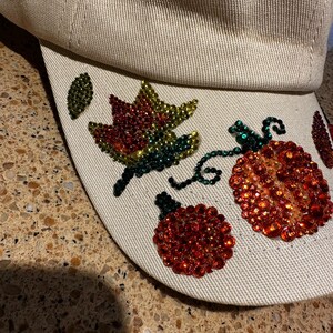Peut inclure: Une casquette de baseball beige avec un motif sur le thème de l'automne représentant une feuille verte, jaune et rouge et deux citrouilles rouges en strass.