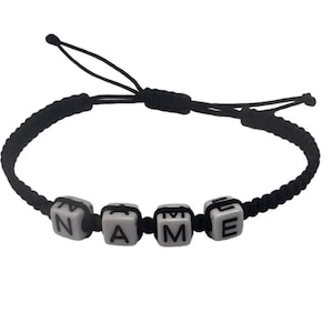 Pode incluir: Pulseira trançada preta com contas quadradas brancas que soletram "NAME". A pulseira ajustável apresenta um fecho de nó deslizante. As letras são em negrito, em preto.