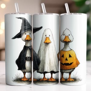 Pode incluir: Três canecas isoladas brancas com palhinhas transparentes. Cada caneca apresenta uma ilustração fantasiosa de um pato com uma fantasia de Halloween: uma bruxa, um fantasma e uma abóbora. Os patos estão sobre um pedaço de grama.