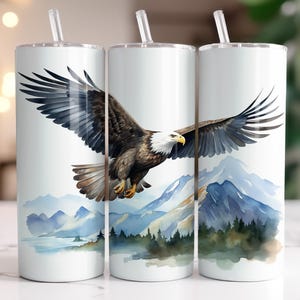 Może przedstawiać: Trzy kubki ze stali nierdzewnej z akwarelowym wzorem przedstawiającym orła bielika lecącego nad pasmem górskim. Orzeł jest brązowy i biały z żółtym dziobem. Góry są niebieskie i zielone na białym tle.
