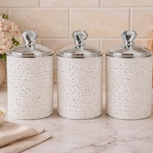Può includere: Tre barattoli cilindrici bianchi con coperchi argentati e finiture a forma di cristallo. I barattoli hanno una superficie strutturata e perlata. Una ciotola di noci, un vaso di fiori e utensili di legno sono sullo sfondo.