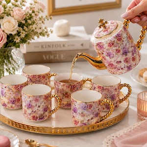 Op de afbeelding: Een porseleinen theepot en vijf bijpassende mokken versierd met een bloemenpatroon van roze en paarse rozen. De theepot en mokken hebben goudkleurige randen en goudkleurige kralenhandvatten. De theepot heeft een goudkleurige tuit en een deksel.