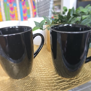 May include: Two black ceramic mugs with rounded handles. The mugs are sitting on a gold-coloured surface.