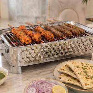 Pode incluir: Uma grelha de mesa em aço inoxidável com espetos de carne grelhada. A grelha está sobre uma superfície de mármore, com pratos de pão naan, cebolas fatiadas e um molho verde. O vapor sobe da grelha quente.