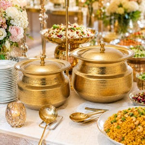 May include: Two round, hammered gold-tone metal serving pots with matching lids, positioned on a table alongside a gold-tone ladle, a small glass candle holder, stacks of white plates, and various bowls filled with prepared food dishes.