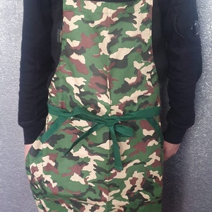 May include: A green, brown and beige camouflage apron with green adjustable straps and a green tie. The apron has a large pocket on the front.