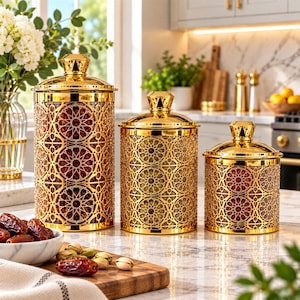 May include: Three cylindrical canisters of varying heights with reflective gold-tone metal lids and a gold-tone lattice overlay featuring a geometric star pattern. The canisters have a clear glass body that reveals the contents inside each container.