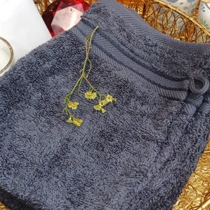 Puede incluir: Una toalla esponjosa de color gris oscuro con un lazo decorativo, colocada en una cesta de alambre dorada con una pequeña rama de flores amarillas.