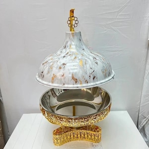 Könnte beinhalten: Ein Speisenwärmer mit einem weiß-goldenen, marmorierten Kuppeldeckel und einem goldenen Knauf. Die Basis besteht aus einer polierten, silberfarbenen Schale in einem verzierten, goldfarbenen Metallständer mit filigranen Ausschnitten.