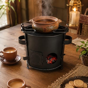 Puede incluir: Una estufa de carbón cilíndrica de metal negro con una puerta lateral y una abertura circular que muestra brasas rojas brillantes. Una olla de barro marrón humeante con un patrón floral descansa encima, junto a dos tazas de barro y galletas.