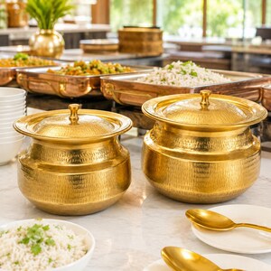 May include: Two round, hammered brass serving pots with matching lids, featuring a wide rim and a rounded body. These metallic vessels are designed for food service and are positioned on a surface alongside white plates and gold-toned spoons.