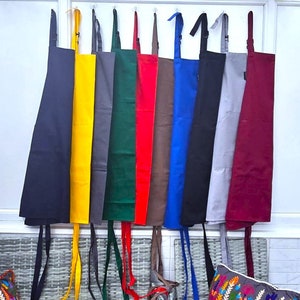May include: Ten different coloured aprons hanging on hooks. The aprons are all made of a plain, solid fabric. The colours are black, yellow, grey, green, red, brown, blue, grey, burgundy, and black.