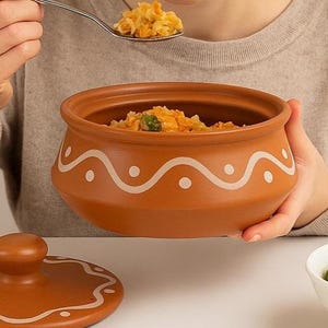 May include: A brown ceramic bowl with a lid, filled with cooked food. The bowl has a white wavy pattern around the rim. A person is holding the bowl and a spoon with food. A white bowl of salad is in the foreground.