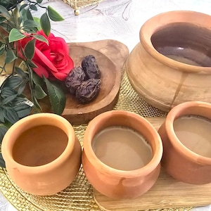 Może przedstawiać: Cztery gliniane kubki z terakoty wypełnione jasnobrązową cieczą, stojące na powierzchni z wzorem w kolorze złotym i brązowym. W tle widać daktyle w drewnianej misce.