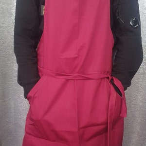May include: A burgundy apron with adjustable straps and a large pocket on the front. The apron has a tie at the waist.