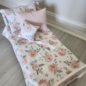 Pode incluir: Uma cama de boneca com estrutura branca e roupa de cama floral. A roupa de cama apresenta um padrão de rosas rosa e pêssego, folhas verdes e pequenas flores sobre um fundo branco. Inclui uma almofada rosa e duas almofadas florais menores.