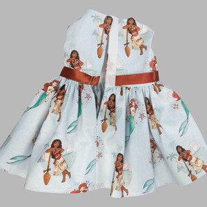 K&ouml;nnte beinhalten: Hellblaues Kleid mit sich wiederholendem Muster einer polynesischen Prinzessin und einer Meerjungfrau. Das Kleid hat ein braunes Band um die Taille und ein wei&szlig;es Band auf dem R&uuml;cken. Das Kleid ist &auml;rmellos und hat einen ausgestellten Rock.