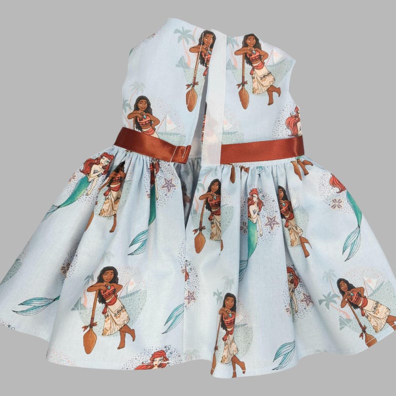 K&ouml;nnte beinhalten: Hellblaues Kleid mit sich wiederholendem Muster einer polynesischen Prinzessin und einer Meerjungfrau. Das Kleid hat ein braunes Band um die Taille und ein wei&szlig;es Band auf dem R&uuml;cken. Das Kleid ist &auml;rmellos und hat einen ausgestellten Rock.
