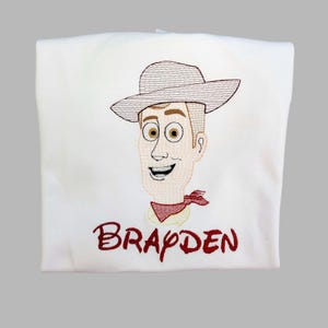 May include: White t-shirt featuring an embroidered cartoon cowboy character with a hat, red neckerchief, and the name "BRAYDEN" in red script. The design is centered on the shirt.