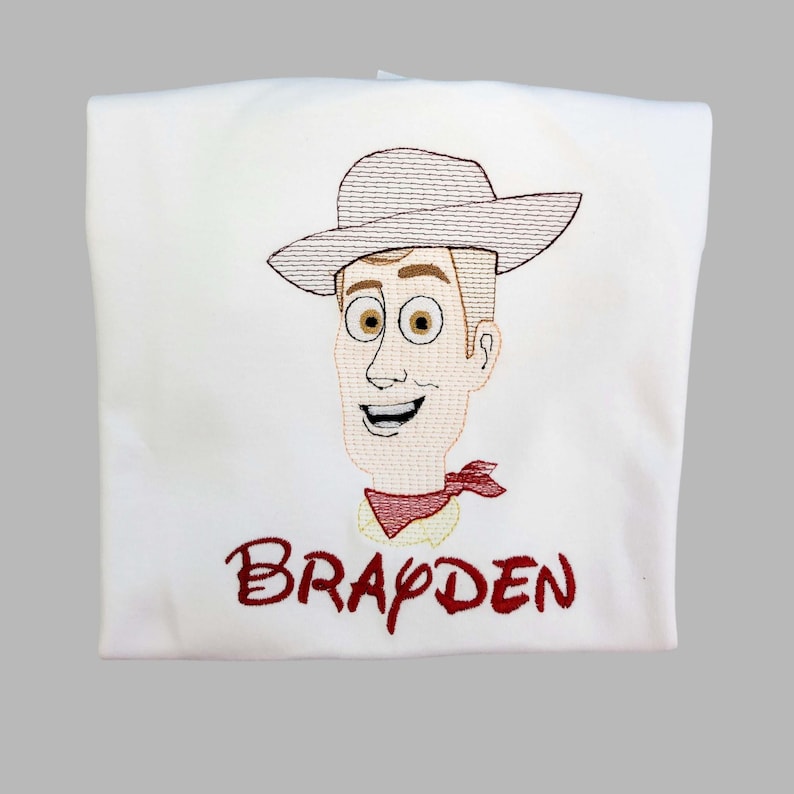 May include: White t-shirt featuring an embroidered cartoon cowboy character with a hat, red neckerchief, and the name "BRAYDEN" in red script. The design is centered on the shirt.