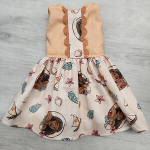 Puede incluir: Vestido de mu&ntilde;eca con corpi&ntilde;o melocot&oacute;n con ribete festoneado y falda estampada. La falda muestra un dise&ntilde;o repetido de un personaje de dibujos animados, veleros, conchas marinas y elementos florales en tonos melocot&oacute;n, azul y rosa.