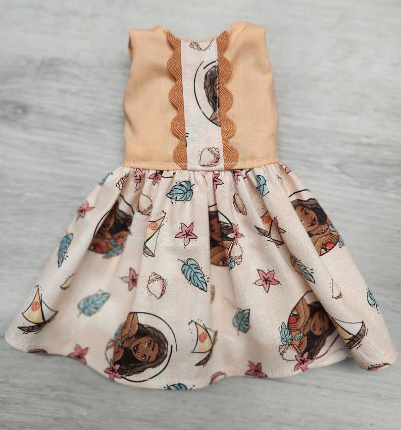 Puede incluir: Vestido de mu&ntilde;eca con corpi&ntilde;o melocot&oacute;n con ribete festoneado y falda estampada. La falda muestra un dise&ntilde;o repetido de un personaje de dibujos animados, veleros, conchas marinas y elementos florales en tonos melocot&oacute;n, azul y rosa.