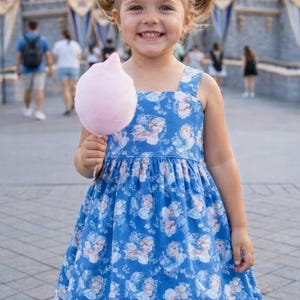 Könnte beinhalten: Ein junges Mädchen lächelt und hält eine rosa Zuckerwatte. Sie trägt ein blaues Kleid mit einem sich wiederholenden Bild einer Prinzessin. Der Hintergrund zeigt einen Vergnügungspark mit einem Schloss.