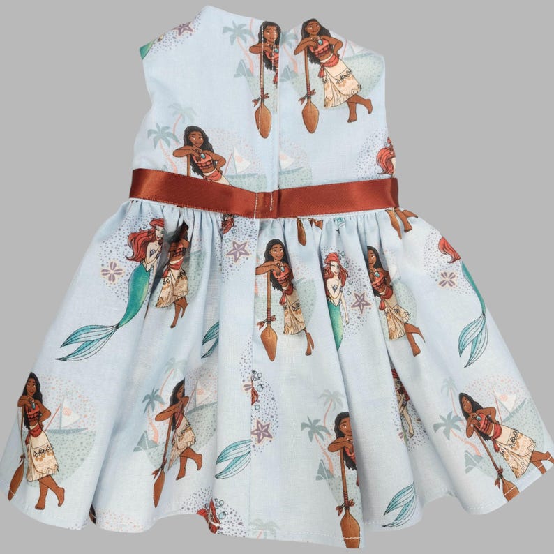 K&ouml;nnte beinhalten: Hellblaues Kleid mit einem sich wiederholenden Muster von Zeichentrickfiguren, darunter eine Meerjungfrau und eine Frau mit einem Paddel. Das Kleid hat ein braunes Band um die Taille und einen Rei&szlig;verschluss hinten. Das Kleid ist f&uuml;r ein Kind.