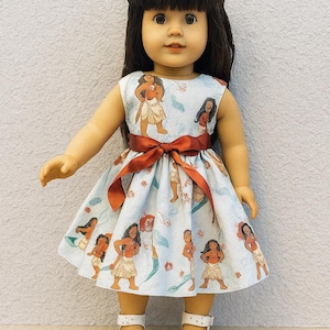 K&ouml;nnte beinhalten: Puppe in einem hellblauen Kleid mit einem sich wiederholenden Druck einer polynesischen Frau. Das Kleid hat eine rote Schleife um die Taille gebunden. Die Puppe tr&auml;gt wei&szlig;e Sandalen.