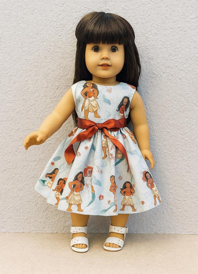 K&ouml;nnte beinhalten: Puppe in einem hellblauen Kleid mit einem sich wiederholenden Druck einer polynesischen Frau. Das Kleid hat eine rote Schleife um die Taille gebunden. Die Puppe tr&auml;gt wei&szlig;e Sandalen.