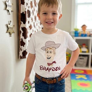 May include: A young child wearing a white t-shirt featuring a cartoon illustration of a cowboy character with a hat and a red bandana. The name "Brapden" is printed below the character. The child is wearing blue jeans and a brown belt.