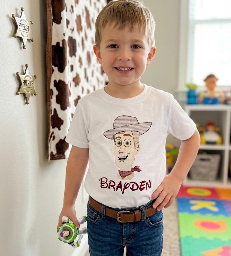 May include: A young child wearing a white t-shirt featuring a cartoon illustration of a cowboy character with a hat and a red bandana. The name "Brapden" is printed below the character. The child is wearing blue jeans and a brown belt.
