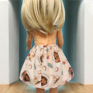 Puede incluir: Una mu&ntilde;eca con una parte superior de color melocot&oacute;n y una falda estampada. La falda presenta ilustraciones de un rostro femenino, veleros y dise&ntilde;os florales sobre un fondo rosa claro. La mu&ntilde;eca tiene el pelo rubio.