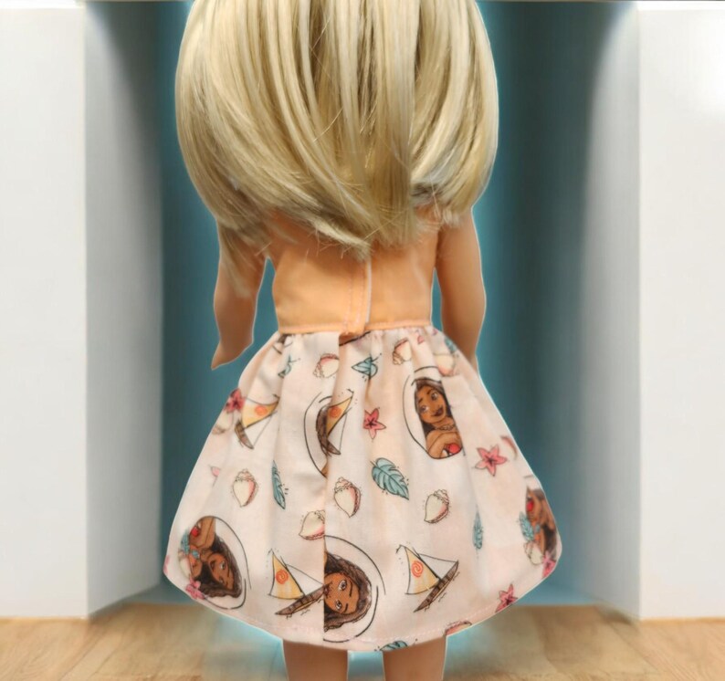 Puede incluir: Una mu&ntilde;eca con una parte superior de color melocot&oacute;n y una falda estampada. La falda presenta ilustraciones de un rostro femenino, veleros y dise&ntilde;os florales sobre un fondo rosa claro. La mu&ntilde;eca tiene el pelo rubio.