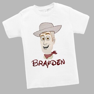 May include: White t-shirt featuring an embroidered cartoon cowboy character with a hat, red neckerchief, and the name "Brayden" in maroon script. The design is centered on the front of the short-sleeved shirt.