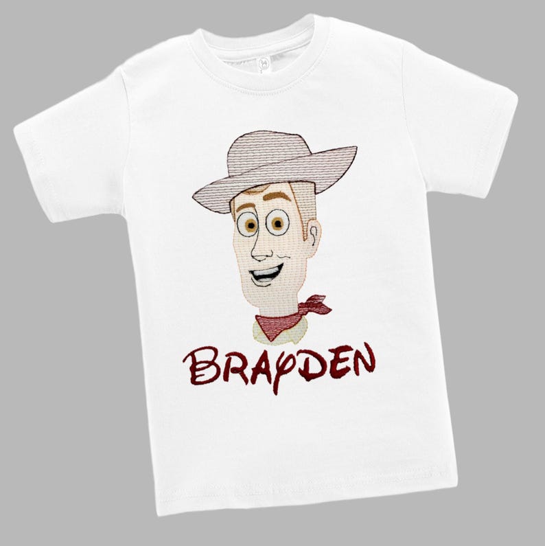 May include: White t-shirt featuring an embroidered cartoon cowboy character with a hat, red neckerchief, and the name "Brayden" in maroon script. The design is centered on the front of the short-sleeved shirt.