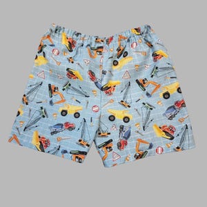 May include: Light blue shorts with an elastic waistband, patterned with various construction vehicles in yellow, orange, red, and blue. The design includes trucks, cranes, and other equipment, set against a grid background.