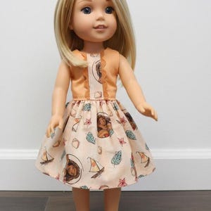 Puede incluir: Mu&ntilde;eca con cabello rubio y ojos azules que lleva un vestido color melocot&oacute;n. El vestido presenta un corpi&ntilde;o con ribete de encaje y una falda estampada con im&aacute;genes de dibujos animados de una mujer, veleros y hojas. La mu&ntilde;eca est&aacute; de pie.