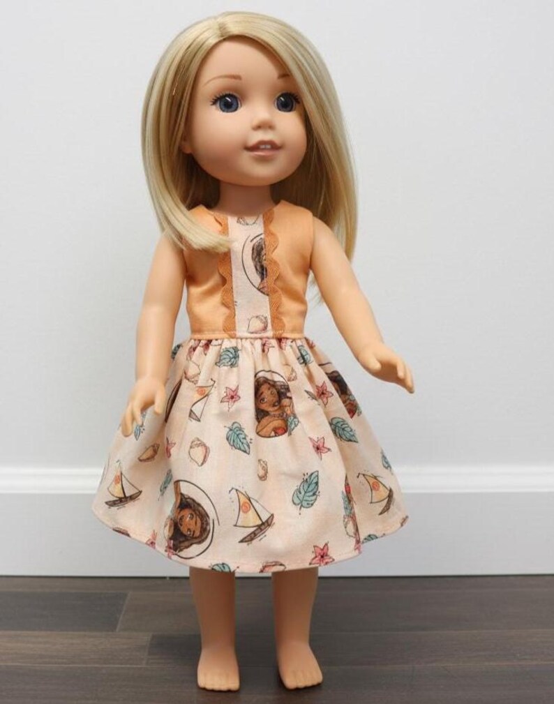 Puede incluir: Mu&ntilde;eca con cabello rubio y ojos azules que lleva un vestido color melocot&oacute;n. El vestido presenta un corpi&ntilde;o con ribete de encaje y una falda estampada con im&aacute;genes de dibujos animados de una mujer, veleros y hojas. La mu&ntilde;eca est&aacute; de pie.