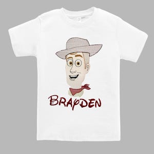May include: White t-shirt featuring an embroidered cartoon cowboy face with a hat and red neckerchief. The name "Brayden" is embroidered in a matching red font below the image. A children's apparel item.