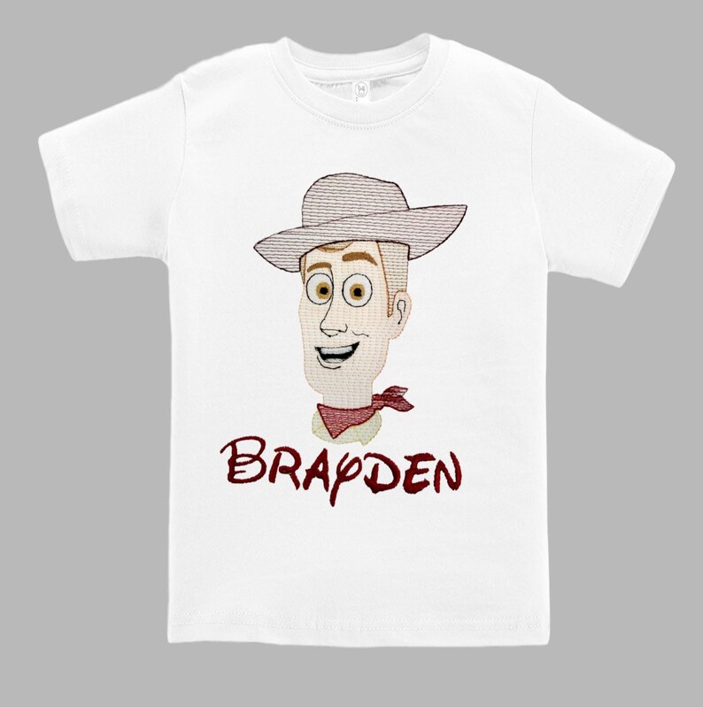 May include: White t-shirt featuring an embroidered cartoon cowboy face with a hat and red neckerchief. The name "Brayden" is embroidered in a matching red font below the image. A children's apparel item.