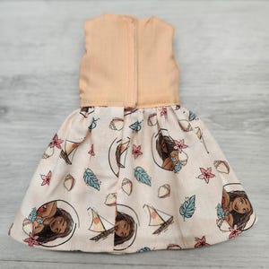 Puede incluir: Un vestido de mu&ntilde;eca con un corpi&ntilde;o color melocot&oacute;n y una falda estampada. La falda presenta un dise&ntilde;o repetido de un retrato de mujer, veleros y motivos florales y de hojas sobre un fondo rosa claro.