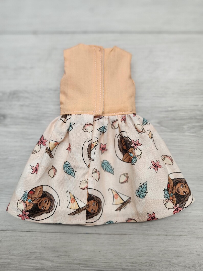Puede incluir: Un vestido de mu&ntilde;eca con un corpi&ntilde;o color melocot&oacute;n y una falda estampada. La falda presenta un dise&ntilde;o repetido de un retrato de mujer, veleros y motivos florales y de hojas sobre un fondo rosa claro.
