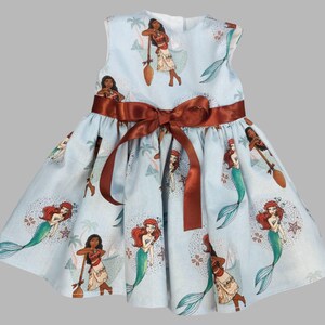 K&ouml;nnte beinhalten: Hellblaues &auml;rmelloses Kleid mit einer braunen Satin-Schleife in der Taille. Das Kleid ist mit einem sich wiederholenden Muster von Zeichentrickfiguren verziert, darunter Meerjungfrauen und eine polynesische Prinzessin. Das Kleid ist f&uuml;r Kinder.
