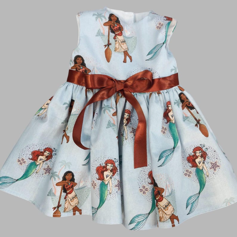 K&ouml;nnte beinhalten: Hellblaues &auml;rmelloses Kleid mit einer braunen Satin-Schleife in der Taille. Das Kleid ist mit einem sich wiederholenden Muster von Zeichentrickfiguren verziert, darunter Meerjungfrauen und eine polynesische Prinzessin. Das Kleid ist f&uuml;r Kinder.