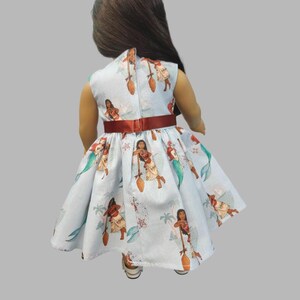 K&ouml;nnte beinhalten: Hellblaues Puppenkleid mit einem Aufdruck einer polynesischen Frau mit Paddel, Meerjungfrauen und tropischen Elementen. Das Kleid hat einen roten G&uuml;rtel und einen hohen Ausschnitt. Die Puppe tr&auml;gt wei&szlig;e Schuhe.
