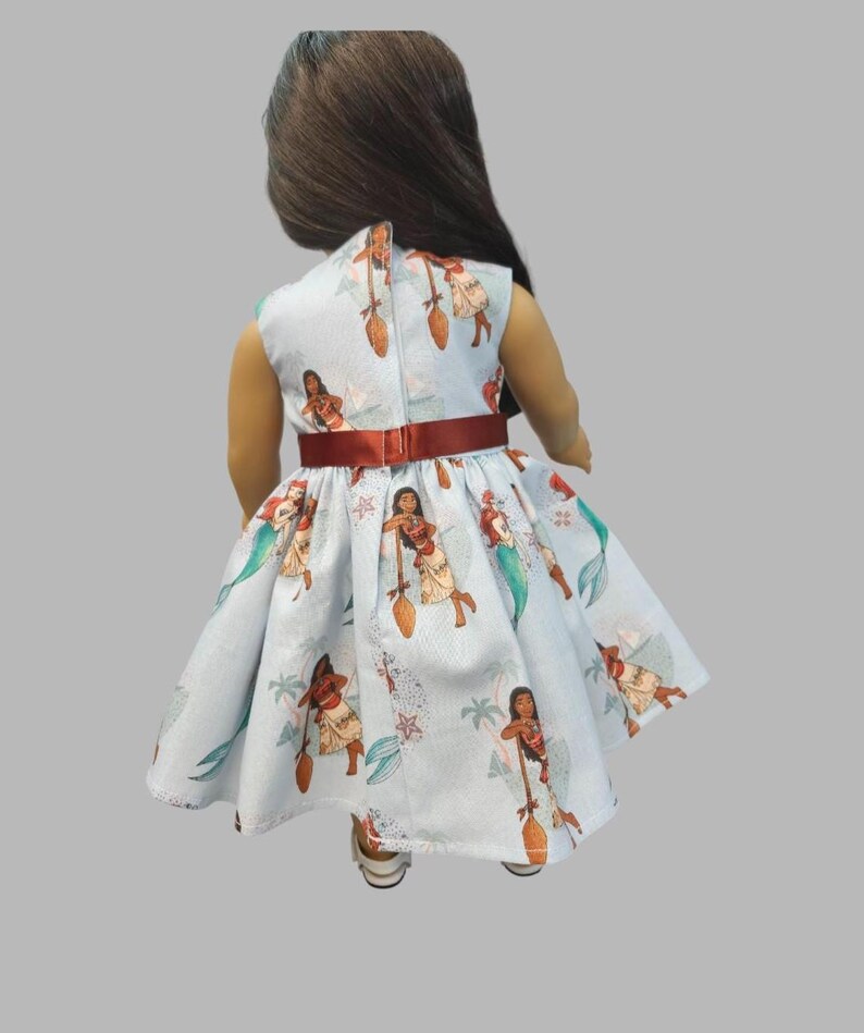 K&ouml;nnte beinhalten: Hellblaues Puppenkleid mit einem Aufdruck einer polynesischen Frau mit Paddel, Meerjungfrauen und tropischen Elementen. Das Kleid hat einen roten G&uuml;rtel und einen hohen Ausschnitt. Die Puppe tr&auml;gt wei&szlig;e Schuhe.