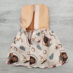 Puede incluir: Un vestido de mu&ntilde;eca con un corpi&ntilde;o color melocot&oacute;n y una falda estampada. La falda muestra un dise&ntilde;o repetido de un personaje de dibujos animados, veleros, conchas marinas y elementos florales sobre un fondo melocot&oacute;n claro. El vestido tiene un cierre de velcro.