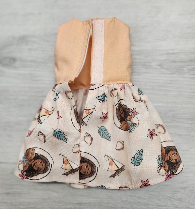 Puede incluir: Un vestido de mu&ntilde;eca con un corpi&ntilde;o color melocot&oacute;n y una falda estampada. La falda muestra un dise&ntilde;o repetido de un personaje de dibujos animados, veleros, conchas marinas y elementos florales sobre un fondo melocot&oacute;n claro. El vestido tiene un cierre de velcro.